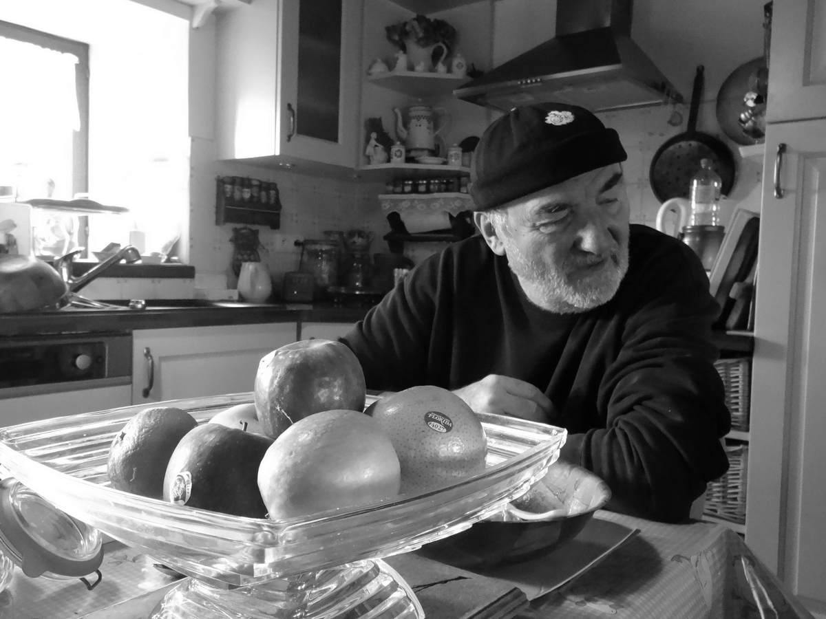 Une cuisine à la campagne une lumière crue du matin. Un homme mûr se détourne du soleil et des fruits.